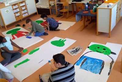 Zu sehen sind Schüler, die auf großen, auf dem Boden liegende Plakate, Olchis malen