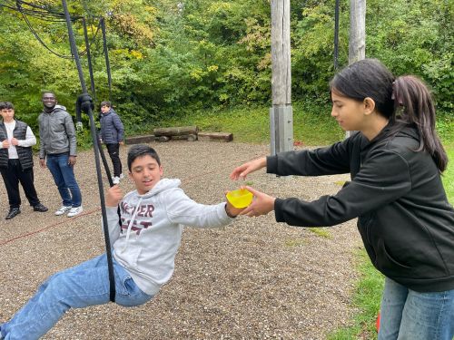 Ein Schüler sitzt auf einem Schaukelseilt. Er übergibt eine Schüssel voll Wasser an eine Schülerin.