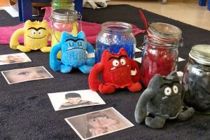Kuscheltiermonster mit verschiedenen Gefühlsausdrücken sitzen auf dem Boden. Daneben liegen Bilder von Kindern mit dem jeweiligen Gefühlsausdruck.