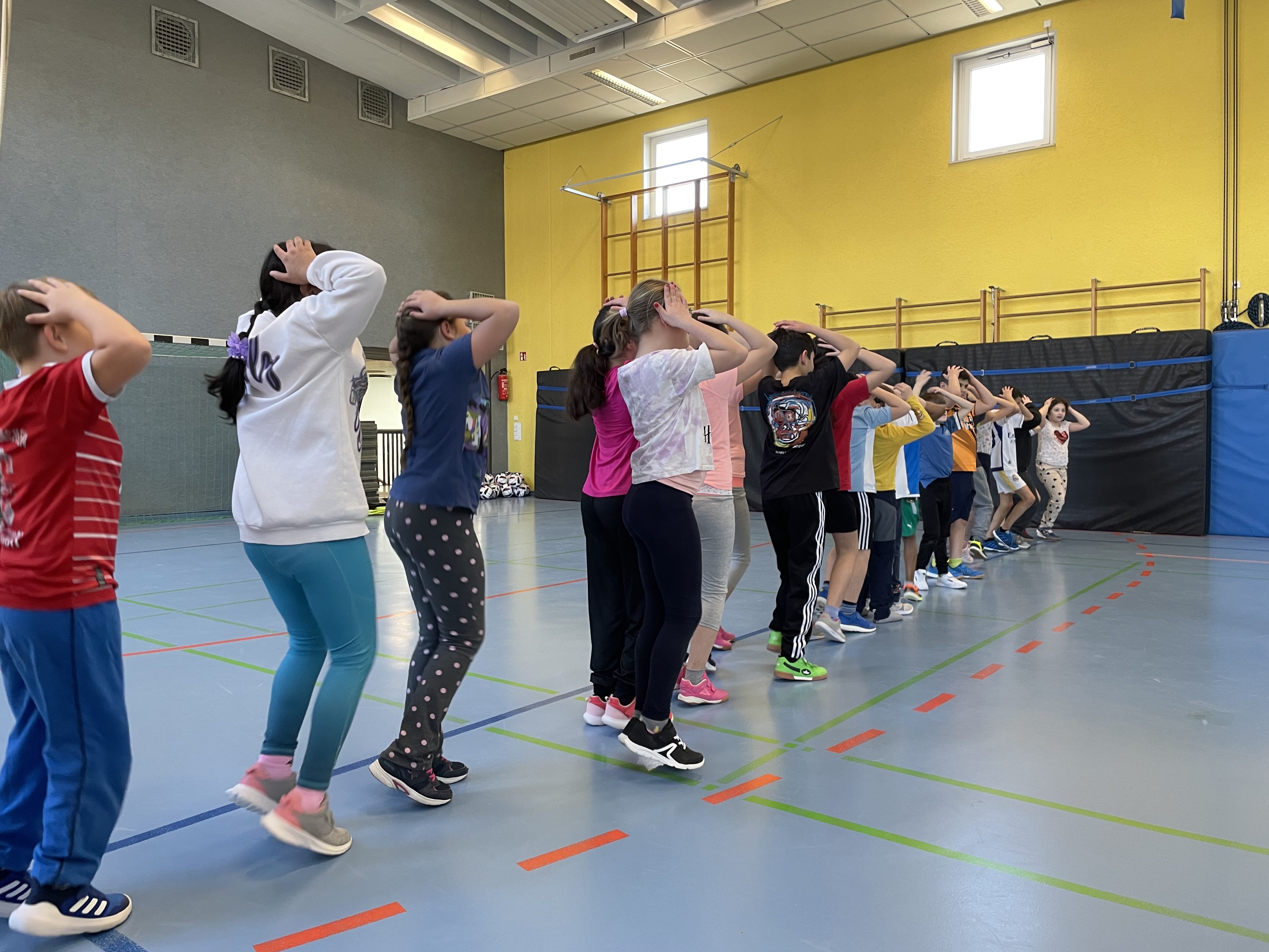 Die Schülerinnen und Schüler stehen in einer Reihe, rennen auf der Stelle und strecken ihre Hände zum Kopf.